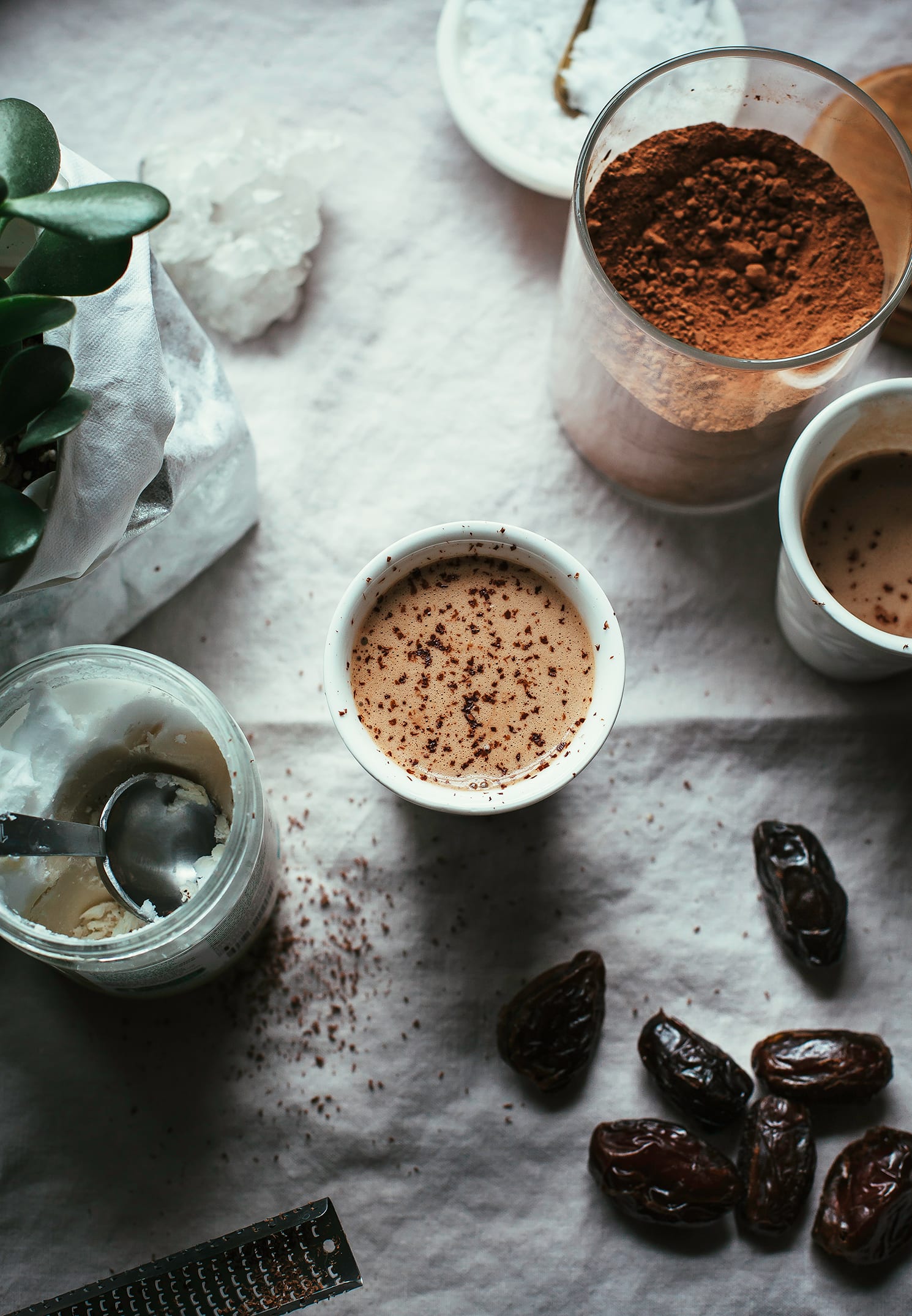 An overhead shot of a cup of hot chocolate dusted with chocolate. To the side is a jar of cacao powder, some dates, and a jar of coconut butter with a measuring spoon inside.