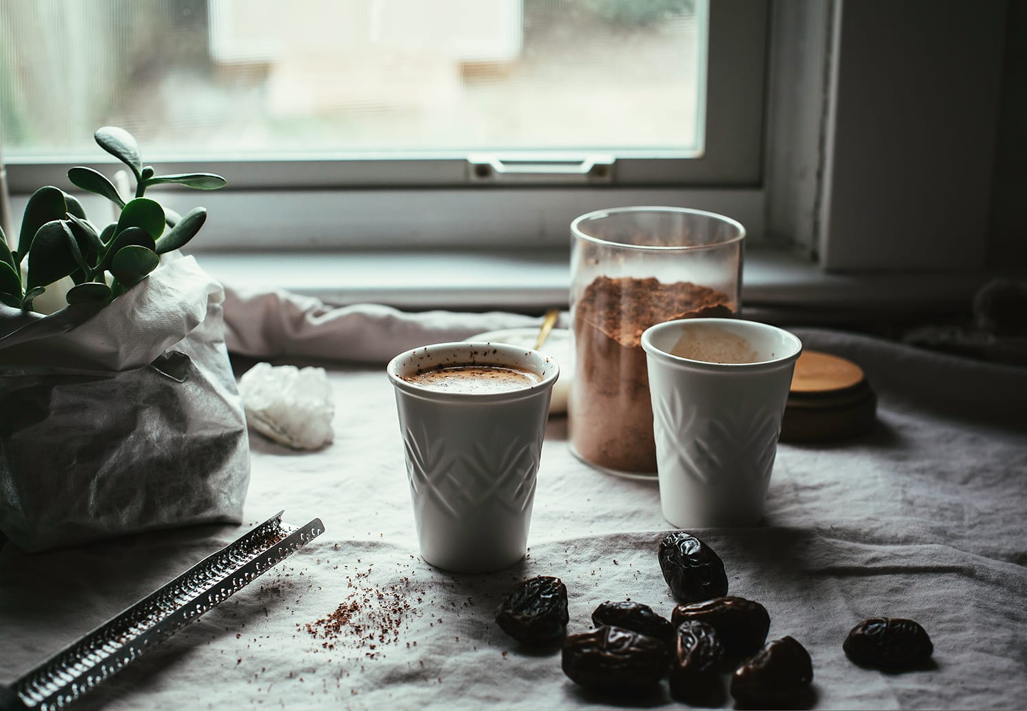 A head on shot of 2 cups with a creamy brown liquid inside. To the side, you see whole dates, a jar of cacao powder, and a plant.