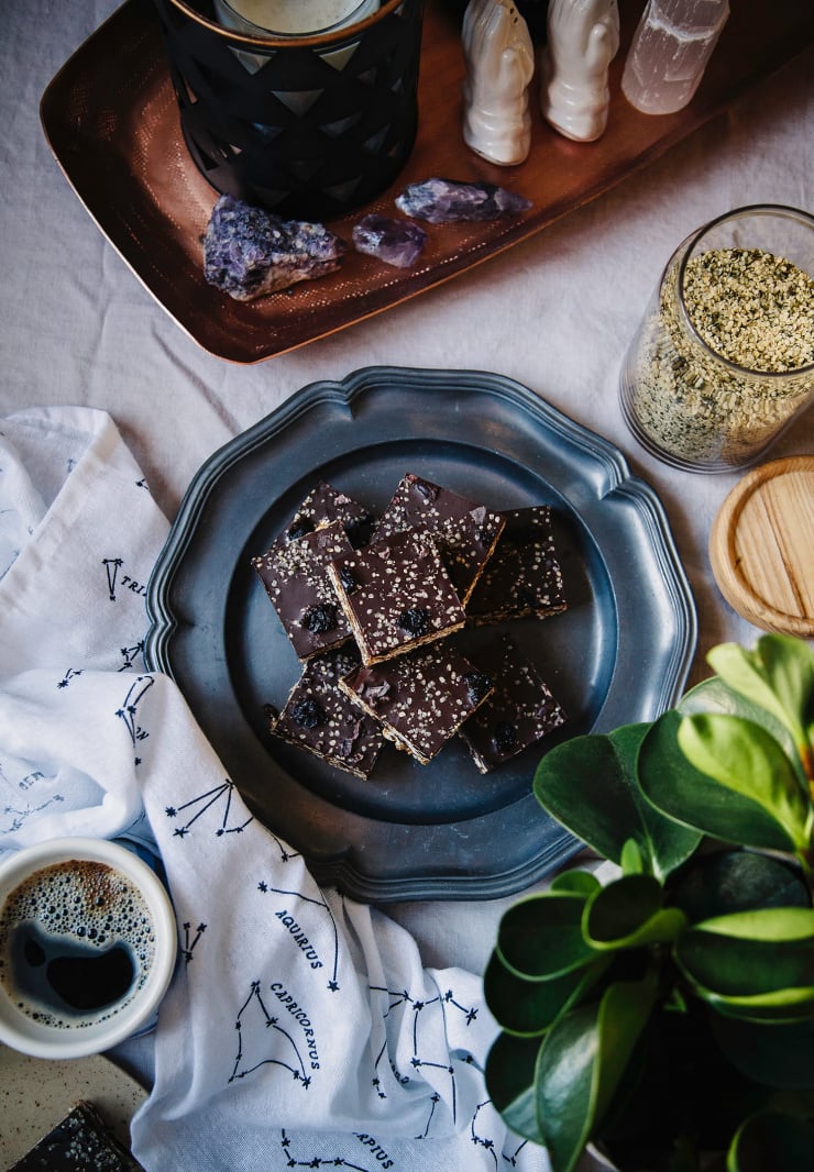 An overhead shot of chocolate covered granola bars on a grey plate. To the side is a jar of hemp seeds, some amethyst crystals, a plant, and a cup of coffee.