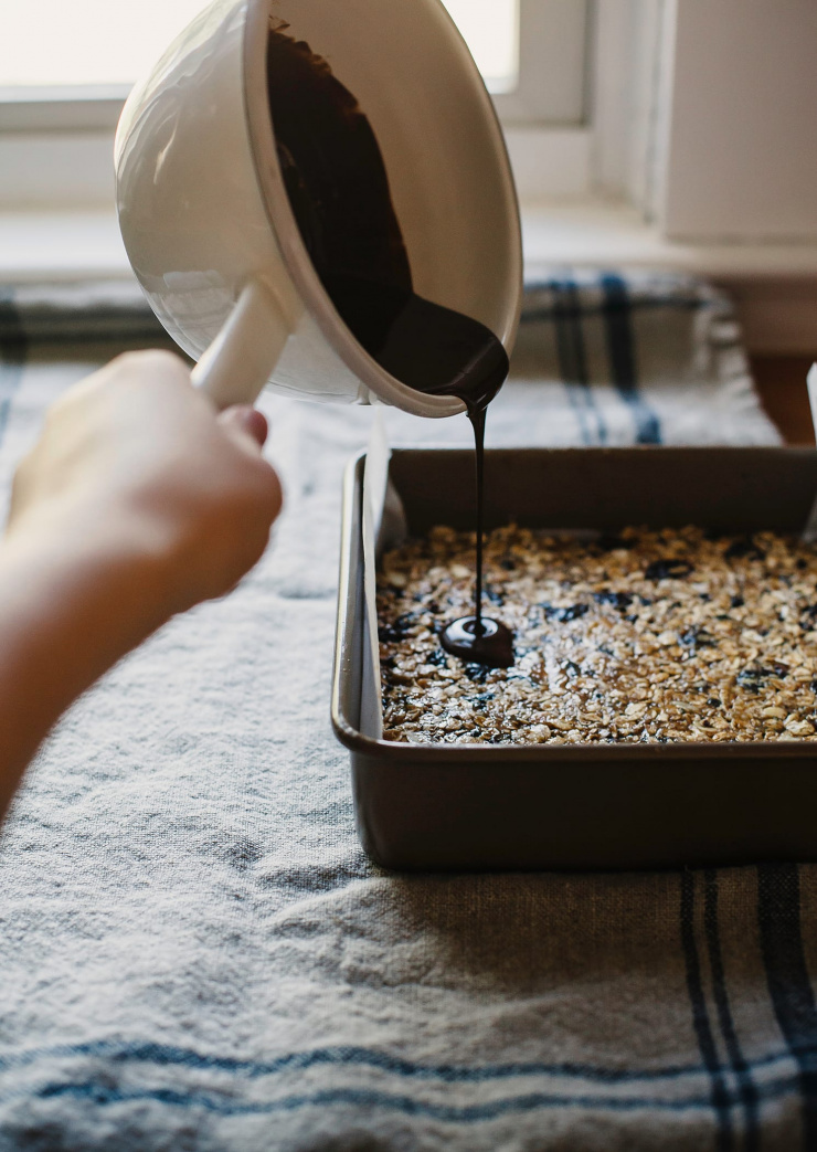 Image shows a hand pouring a saucepan of melted chocolate that is pouring out over a pan of no bake oat bars.