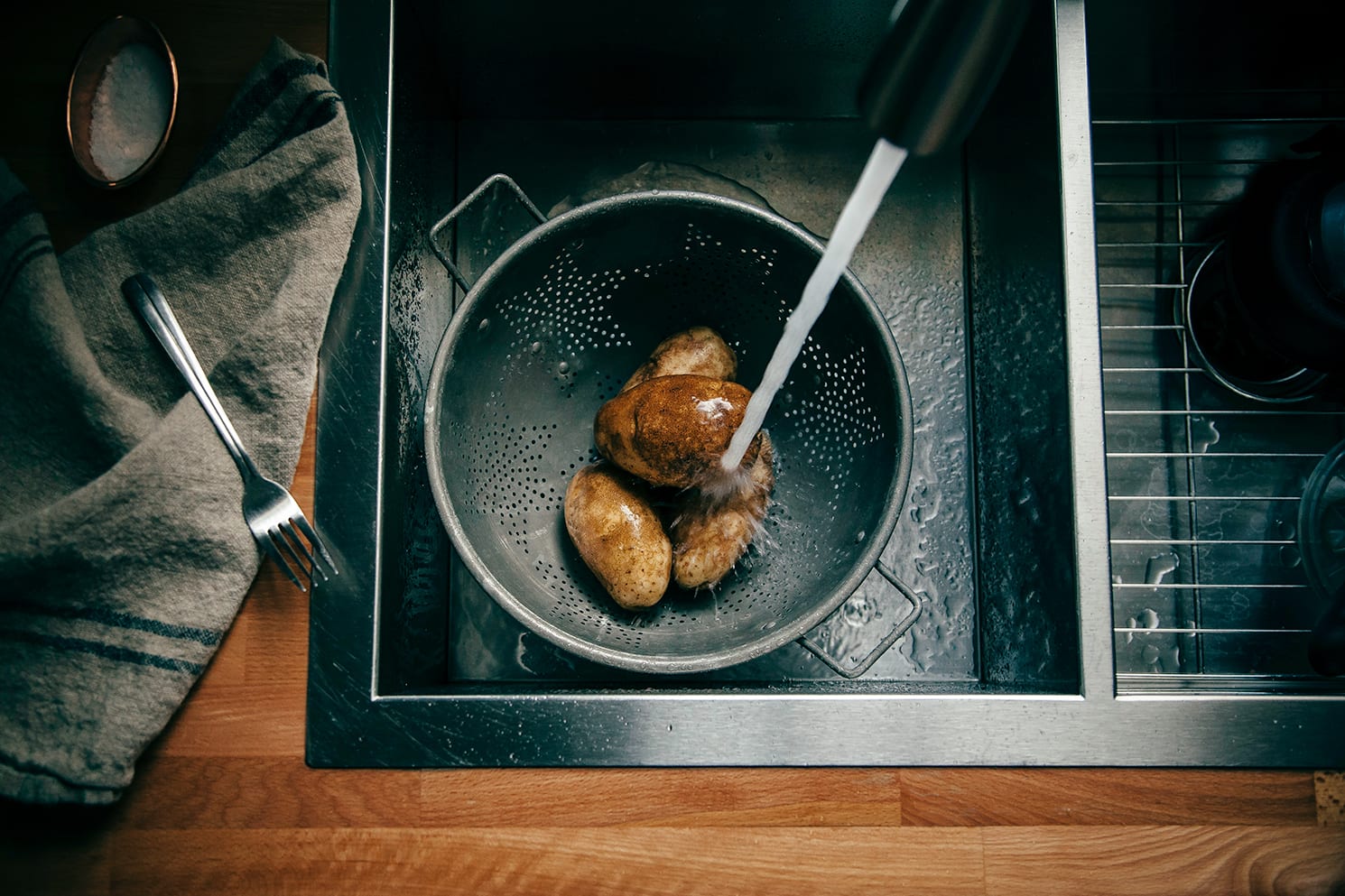 Image shows potatoes in a colander being sprayed by water in a kitchen sink.
