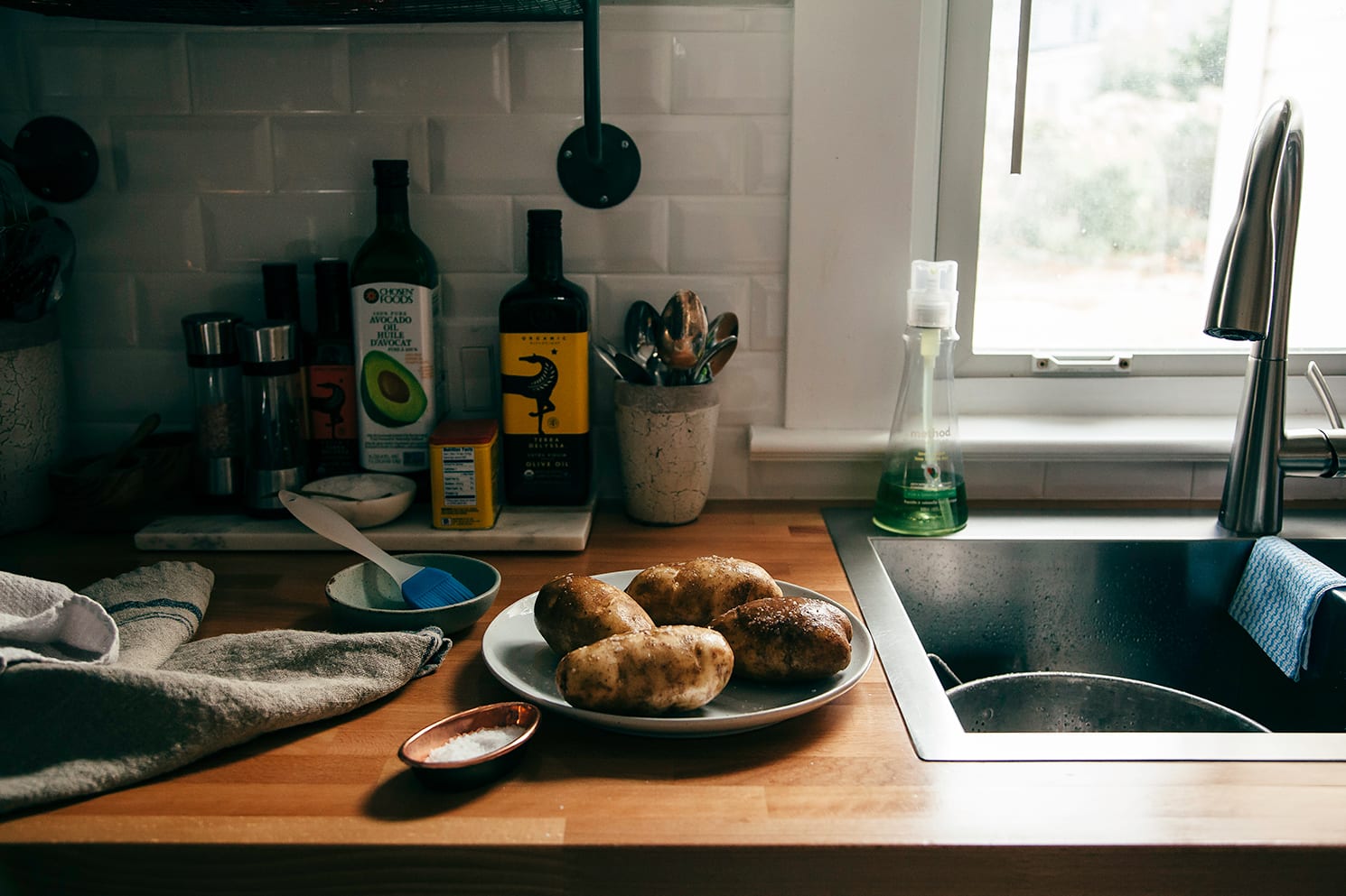 Image shows baking potatoes on a plate on top of a kitchen counter. They are topped with coarse salt.