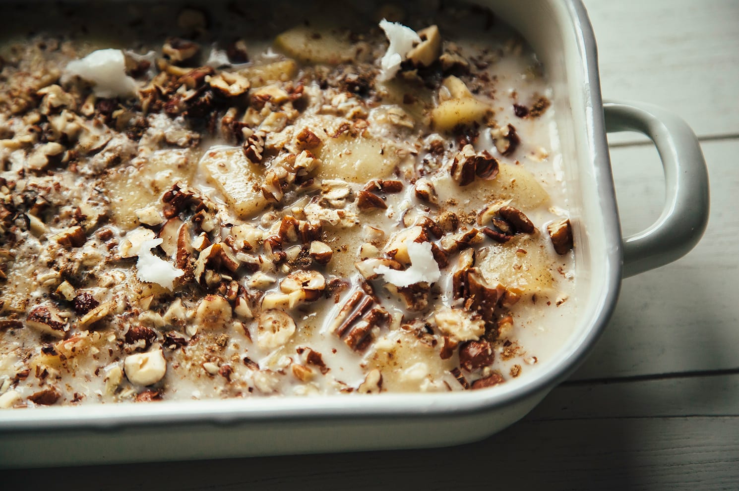 Image shows an assembled apple spice baked oatmeal in a white enamelware baking dish, pre-bake.