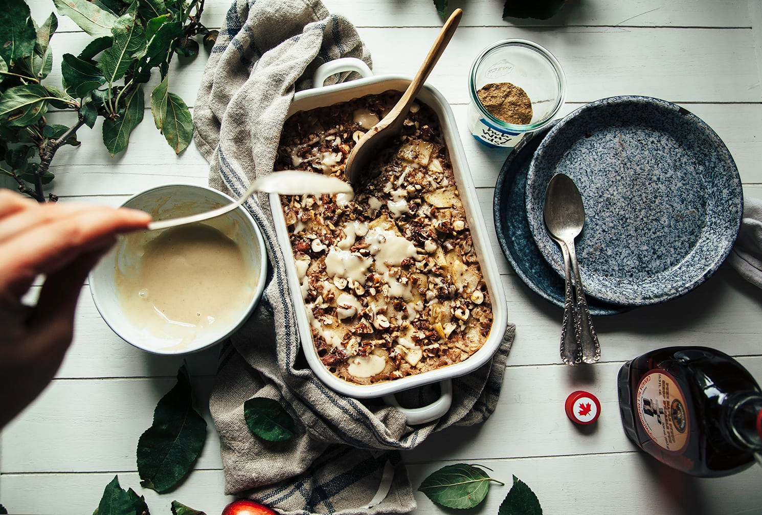 A hand is drizzling a creamy sauce over a dish of baked oatmeal that is topped with cooked apples and nuts.