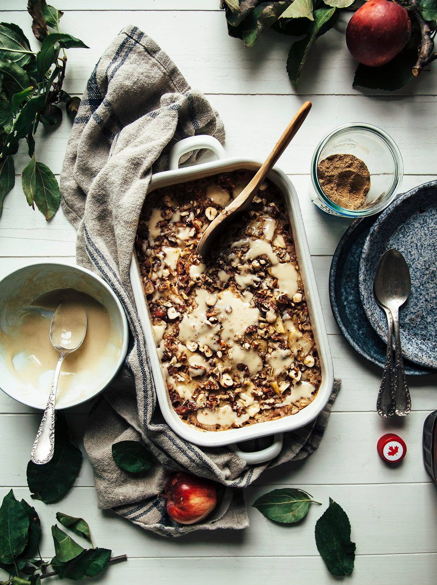 An overhead shot of baked oatmeal that is topped with cooked apples and nuts. A bowl of creamy sauce is seen to the side.