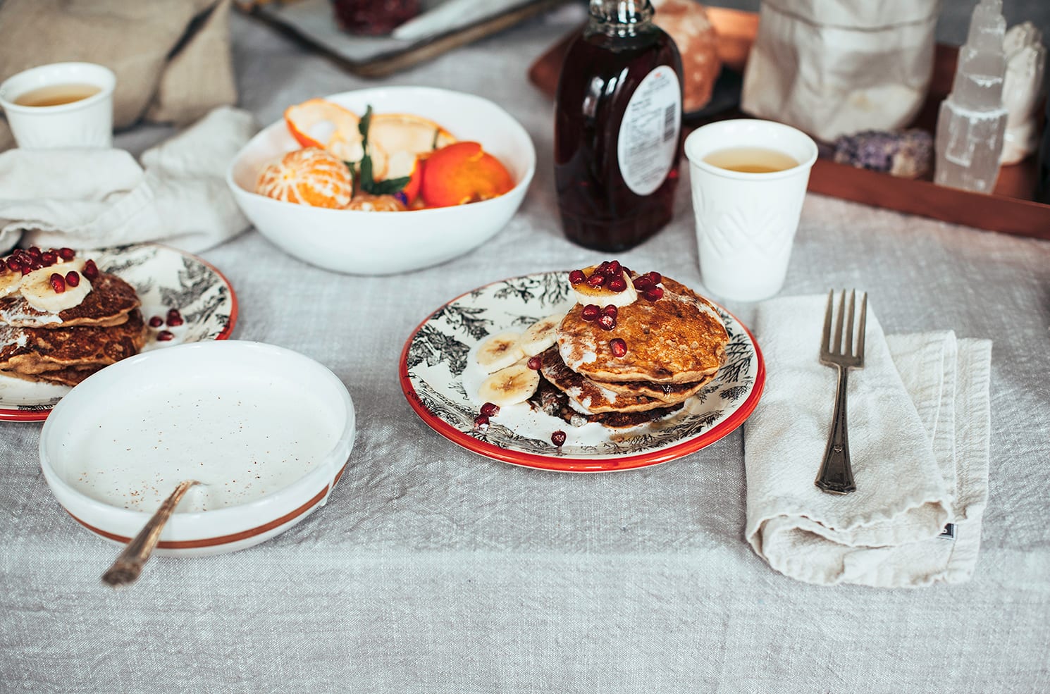 A 3/4 angle image shows a plate of pancakes drizzled with a creamy sauce and topped with sliced banana and pomegranate seeds. A bowl with peeled clementines is also shown to the side.