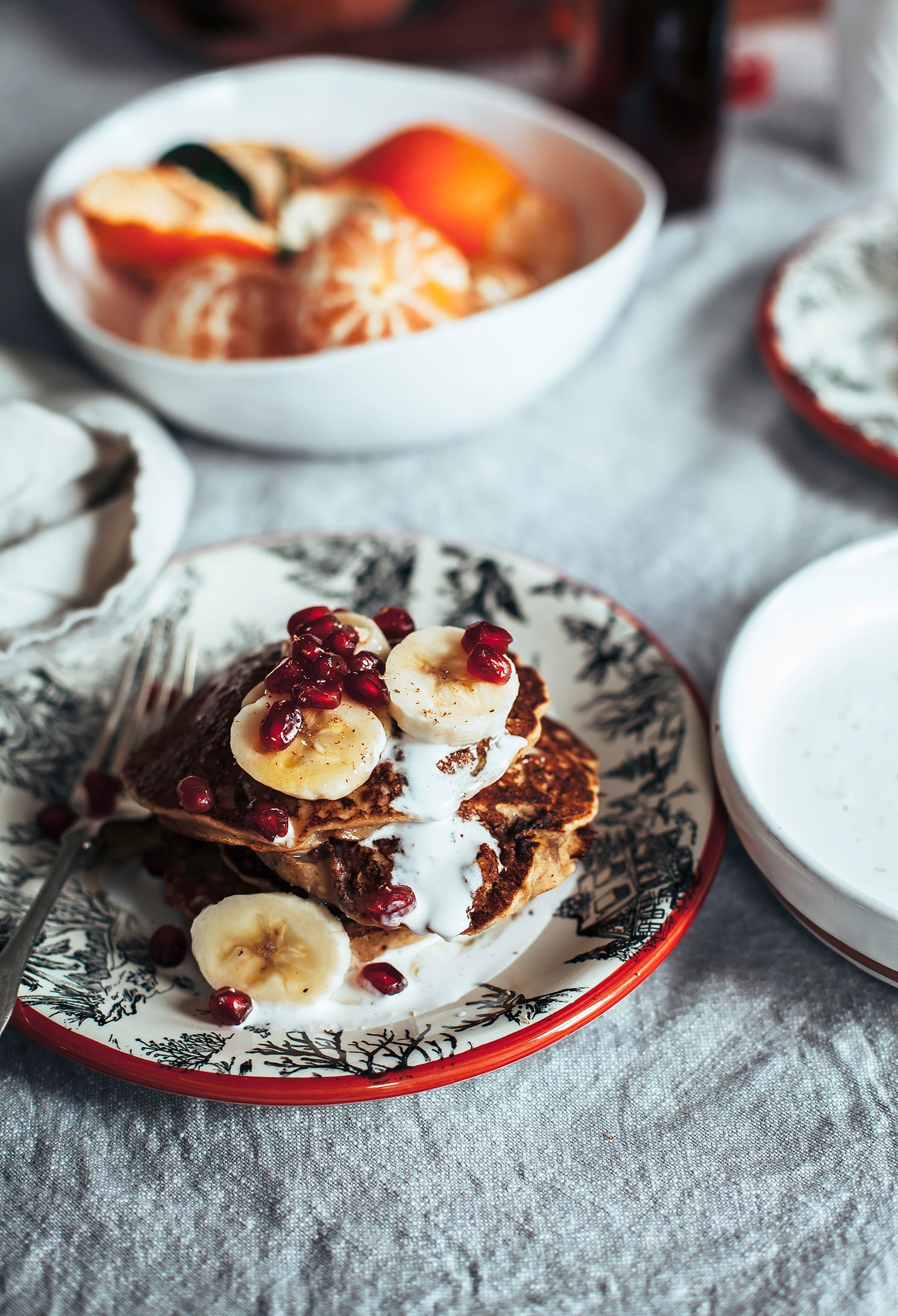 A 3/4 angle image shows a plate of pancakes drizzled with a creamy sauce and topped with sliced banana and pomegranate seeds.