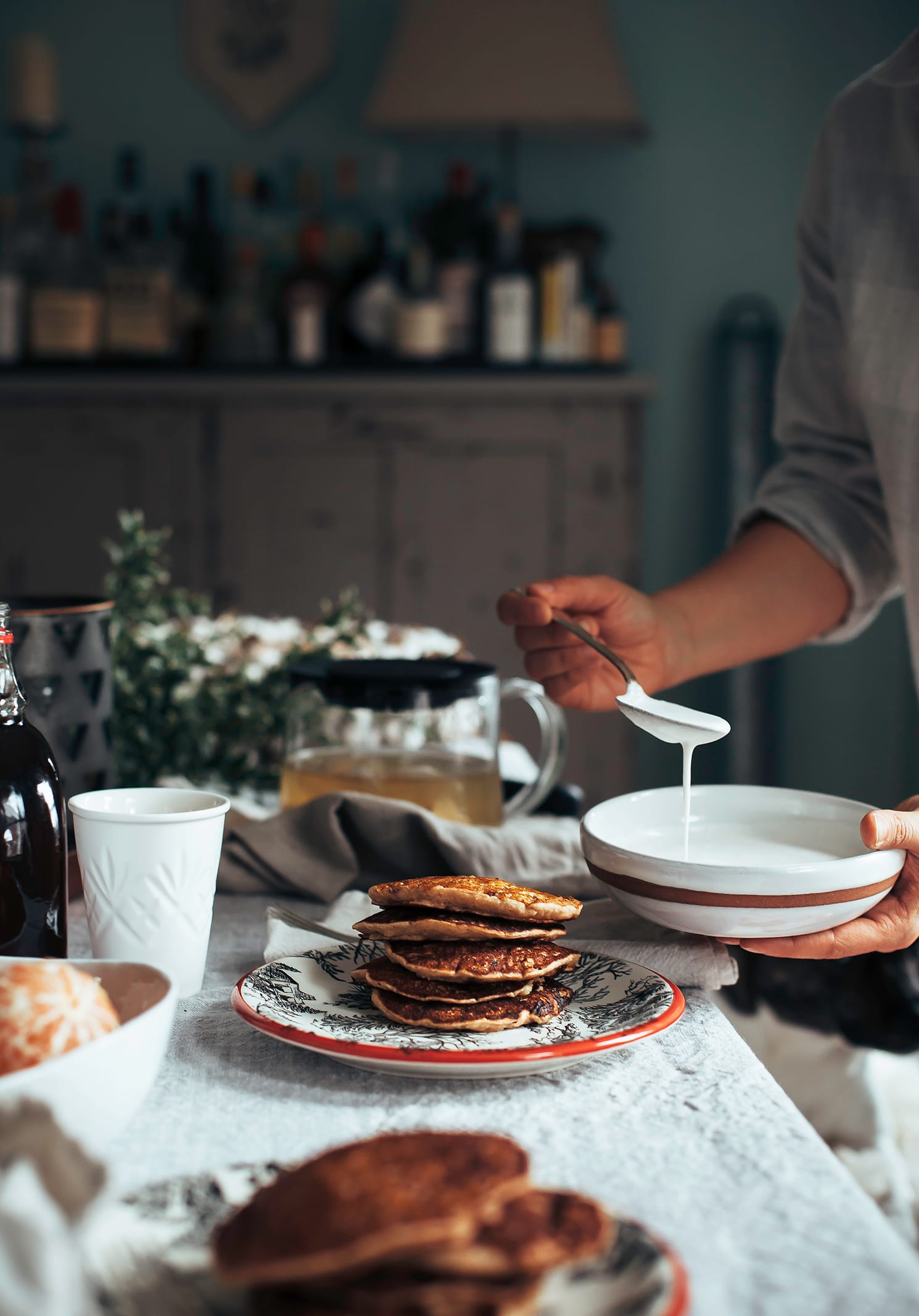 a head-on image shows a set, festive table for brunch and a hand is spooning a creamy sauce to put on a stack of pancakes nearby.