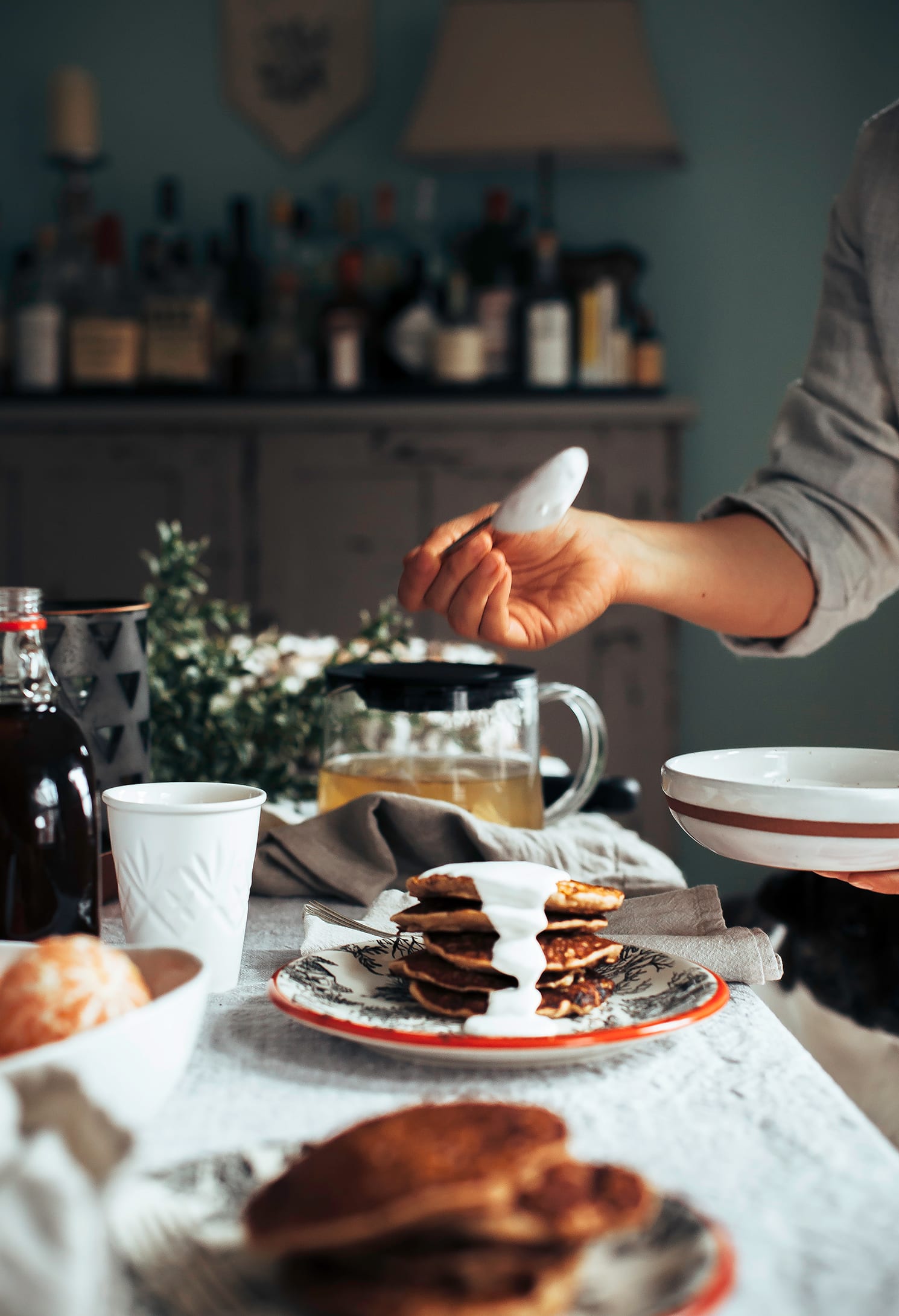 a head-on image shows a set, festive table for brunch and a hand is spooning a creamy sauce to put on a stack of pancakes nearby.