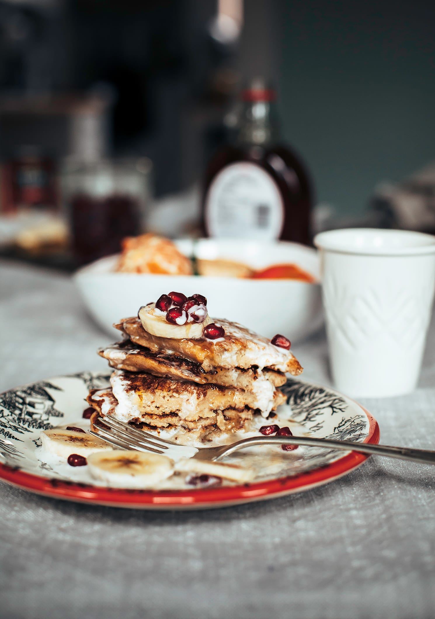 A head-on shot shows a plate of pancakes drizzled with a creamy sauce and topped with sliced banana and pomegranate seeds. The pancakes have been cut into.