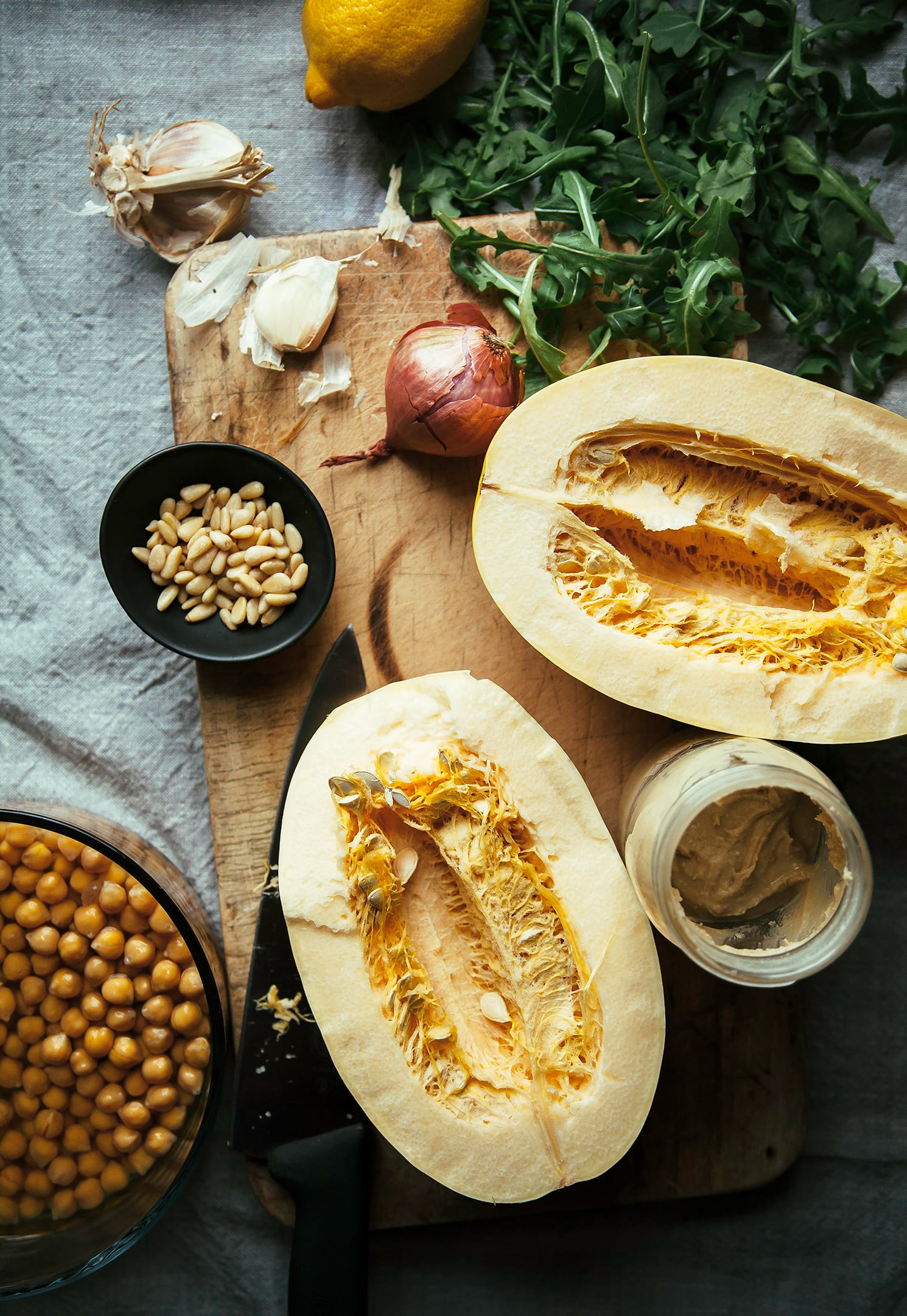 An overhead shot of ingredients for stuffed spaghetti squash on top of a worn wooden cutting board that’s set on top of a grey linen tablecloth.