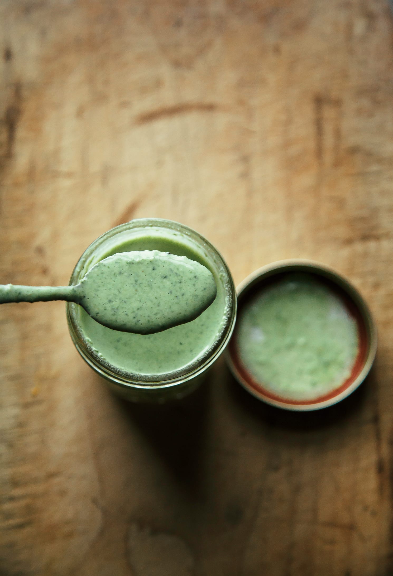 An overhead shot of a jar of bright green garlicky arugula cream sauce in a mason jar. A spoon is lifting out a spoonful of sauce. It is photographed on a worn wood background.