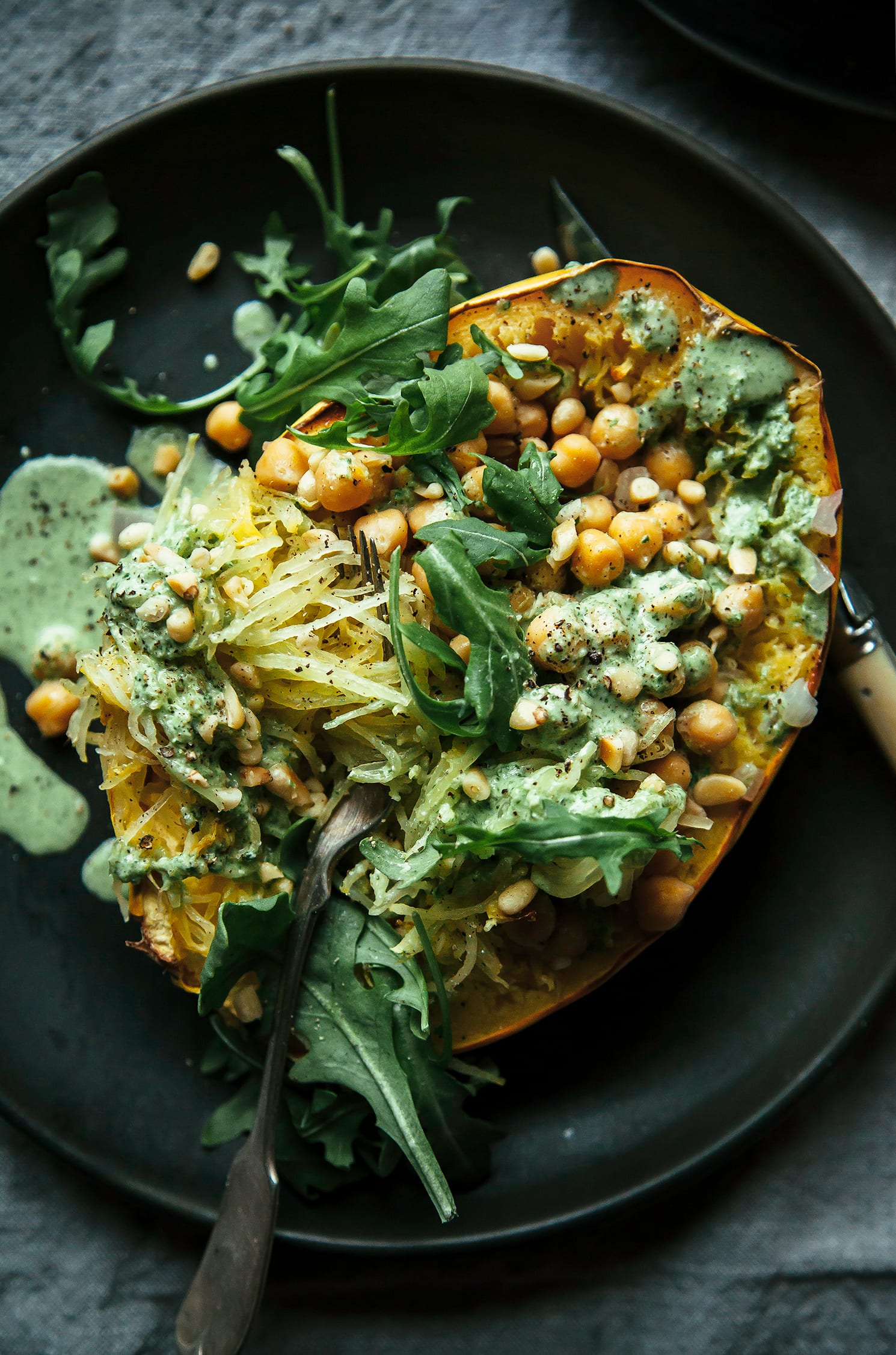 An overhead shot of a stuffed spaghetti squash half on top of a matte black plate that’s set on top of a grey linen tablecloth. Inside the spaghetti squash is chickpeas, bright green garlicky arugula cream and leaves of arugula as well.