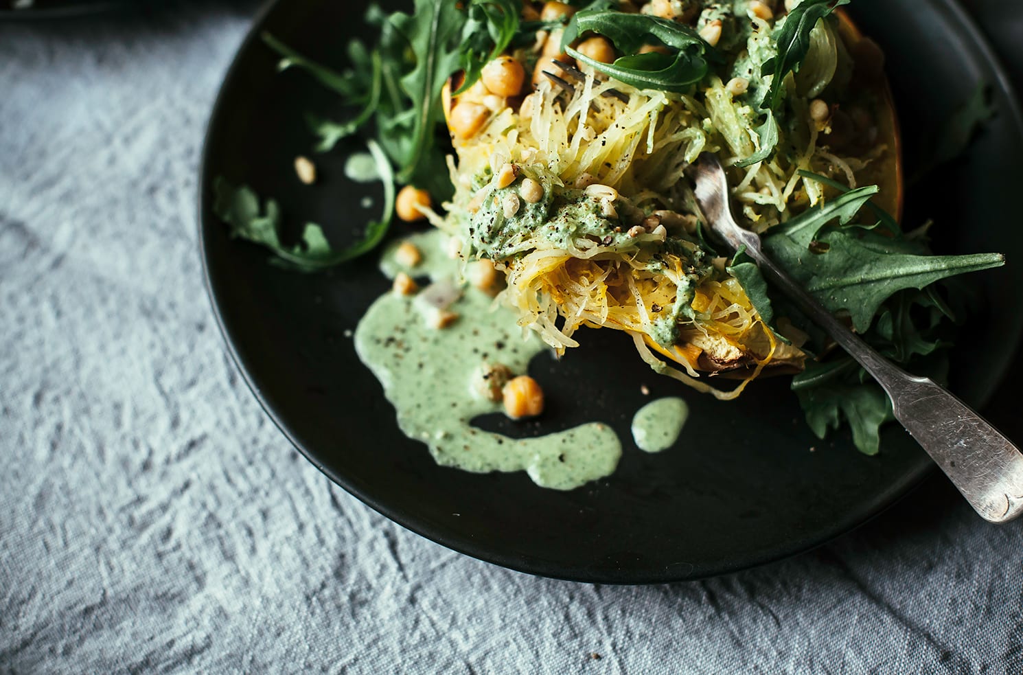 An overhead shot of stuffed spaghetti squash with a bright green arugula cream, leaves of arugula, and chickpeas. The squash is served on a black plate on top of a grey linen tablecloth.