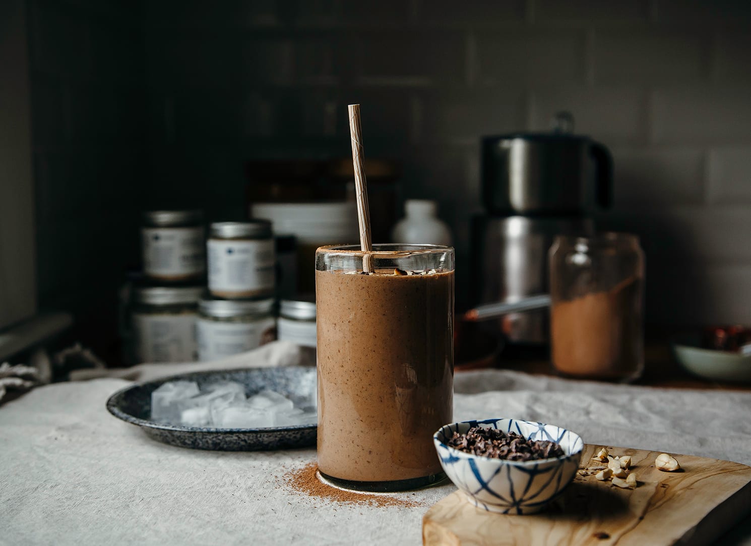 A head-on shot of a double chocolate espresso smoothie that is garnished with chopped hazelnuts and cacao nibs. The smoothie is on top of a beige tablecloth with a small bowl of cacao nibs to the side.