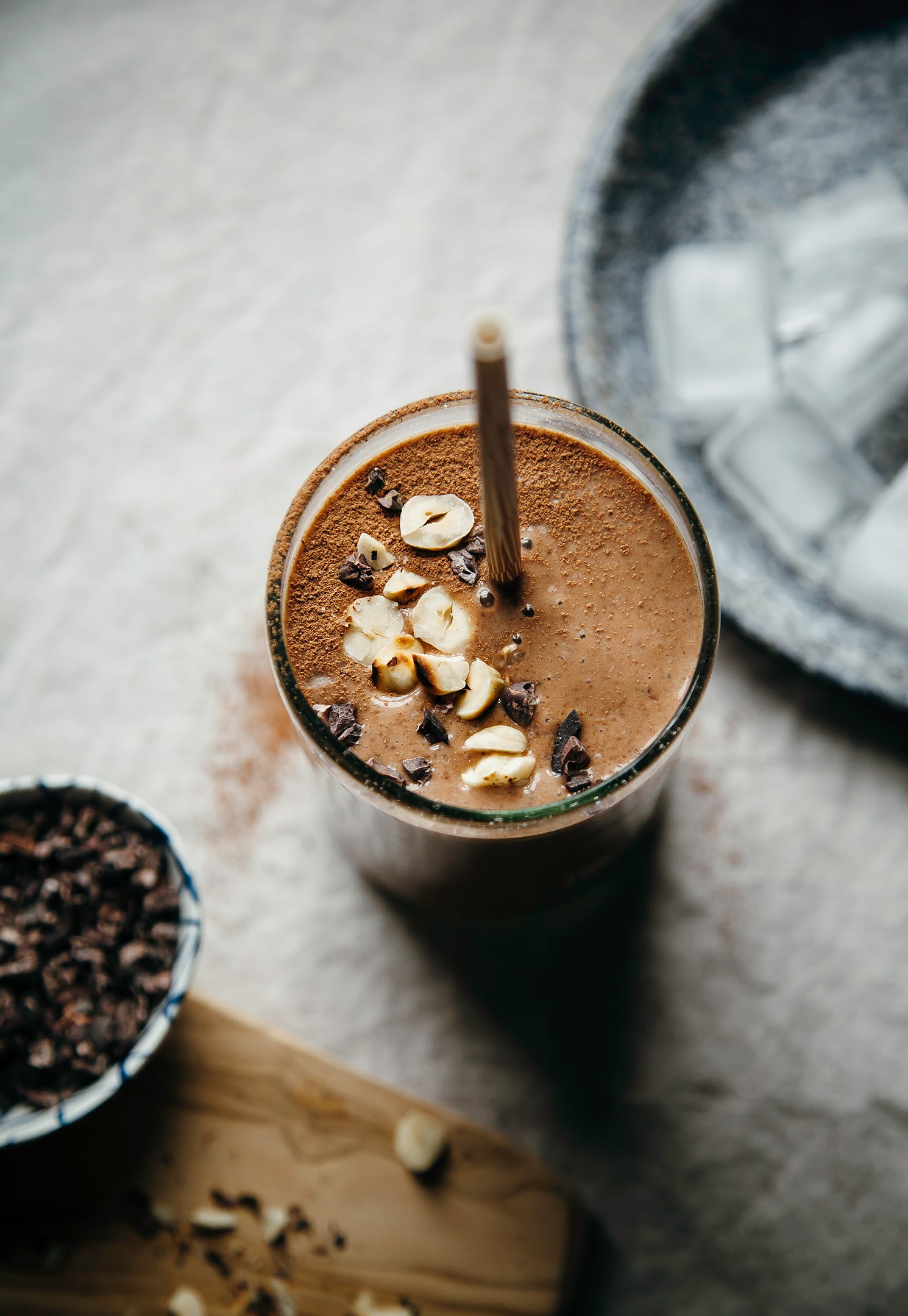 An overhead shot of a double chocolate espresso smoothie that is garnished with chopped hazelnuts and cacao nibs. The smoothie is on top of a beige tablecloth with a small bowl of cacao nibs to the side.