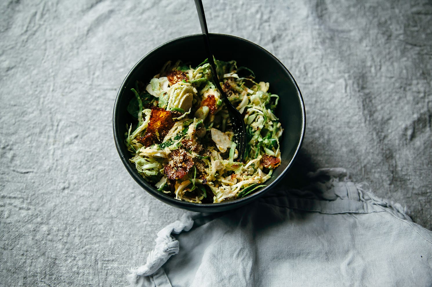 An overhead shot of a small, matte black bowl containing some shaved brussels sprouts and deep red pieces of rice paper "bacon"