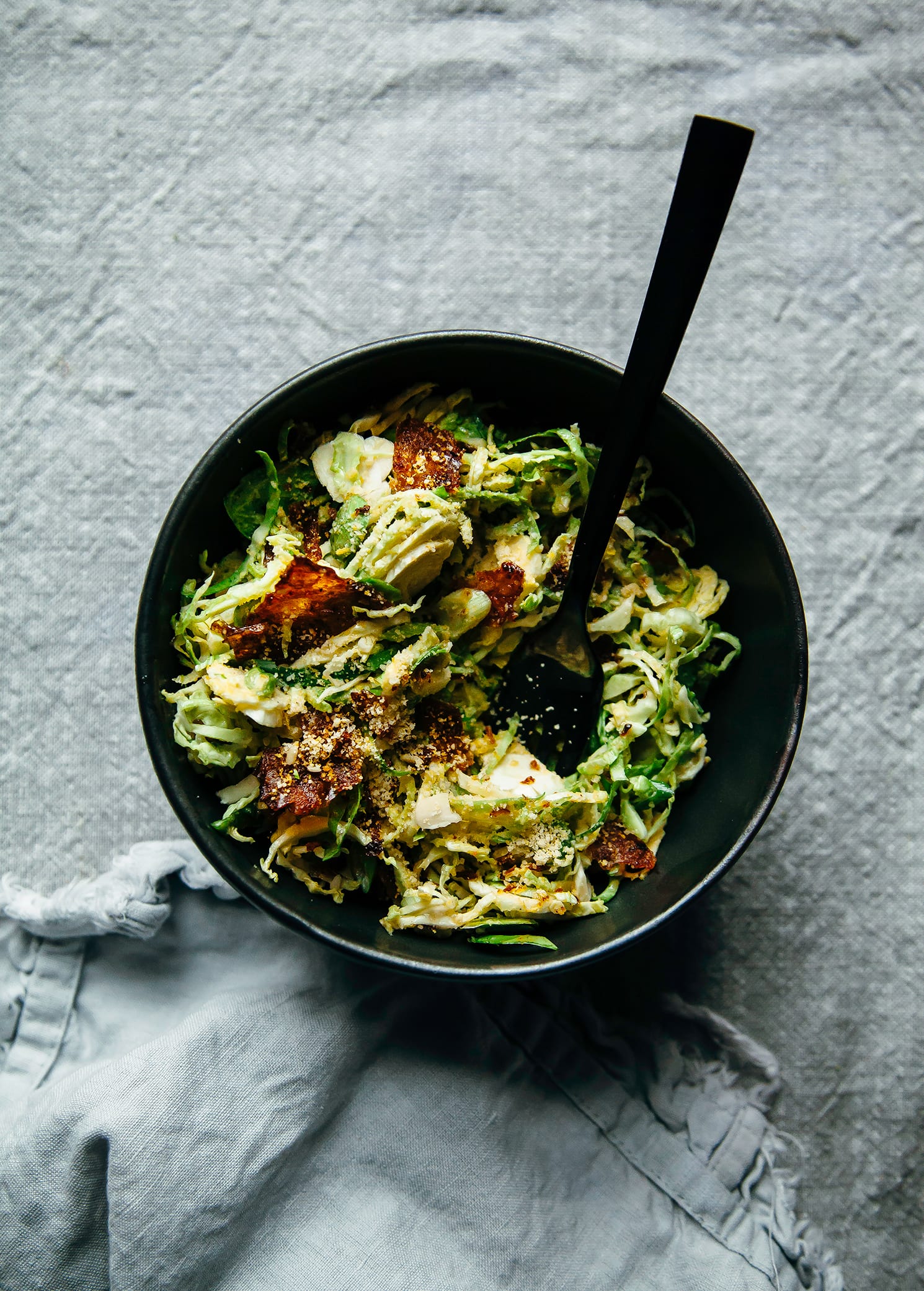 An overhead shot of a small, matte black bowl containing some shaved brussels sprouts and deep red pieces of rice paper "bacon"