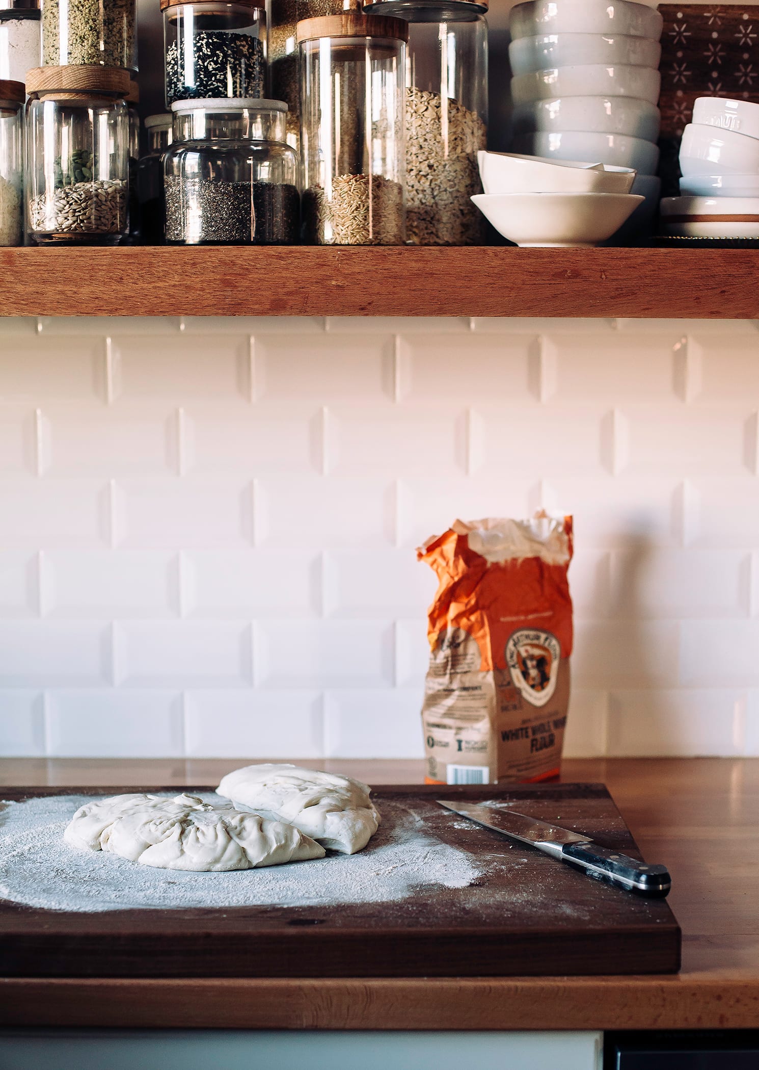 Image shows pizza dough being cut on a cutting board on top of a kitchen counter.