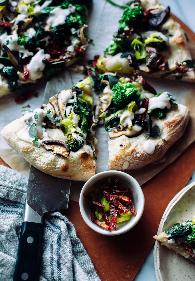 A 3/4 angle shot of a sliced pizza topped with a vegan white sauce, cooked broccoli rabe, shiitake mushrooms, pepperoncini peppers, sun dried tomatoes and oregano.
