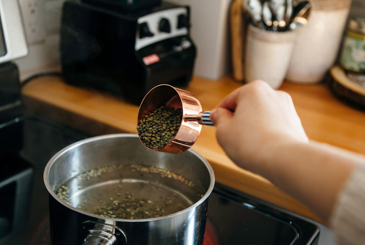 Image shows a hand pouring lentils into a saucepan full of water from a copper measuring cup,