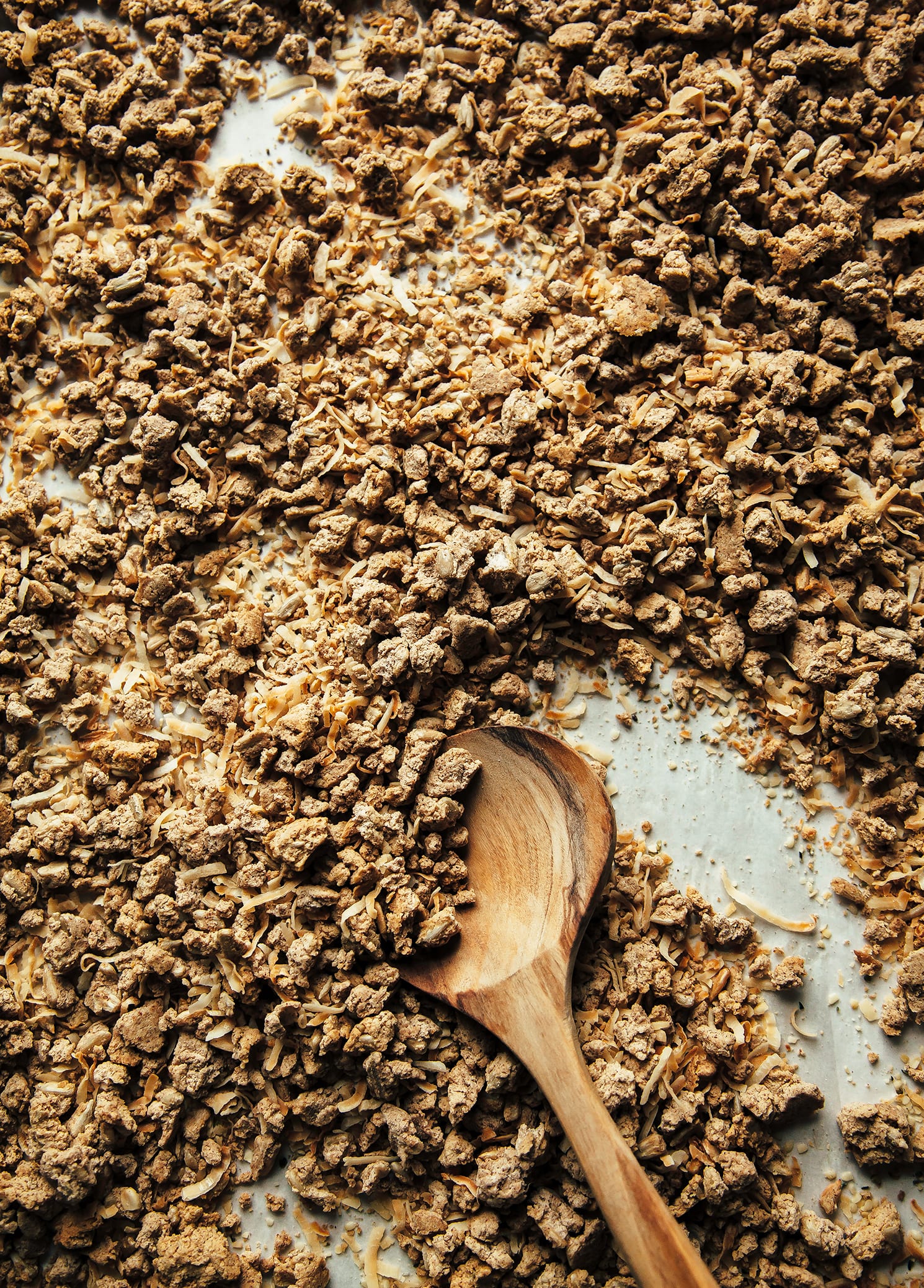 An up close, overhead shot of a tray of granola. A wooden spoon is stirring it up.