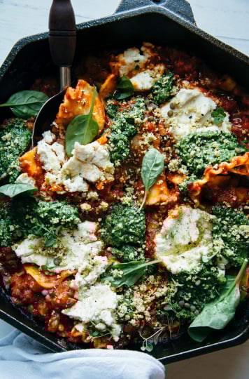 An overhead shot of a rustic, free form skillet lasagna topped with almond "ricotta" dollops, blobs of pesto, and spinach leaves. The dish is photographed in an octagonal cast iron pan.
