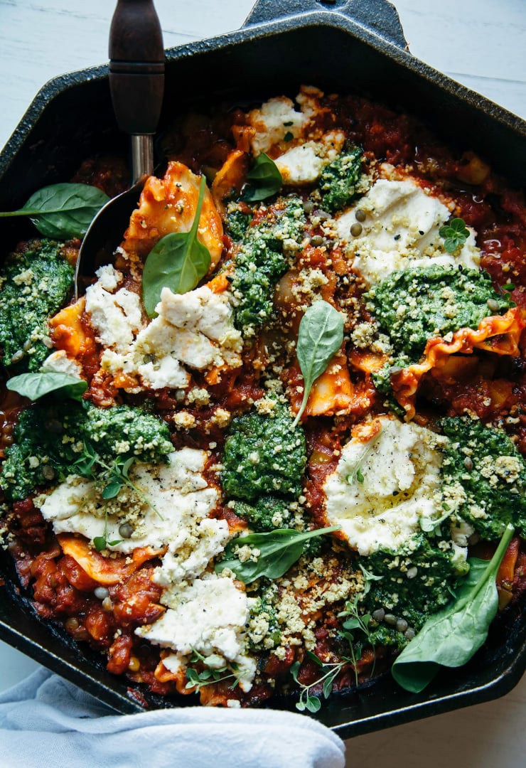 An up close, overhead shot of a rustic skillet lasagga topped with creamy white dollops of vegan "ricotta" and pesto. Part of a roundup of lentil recipes.