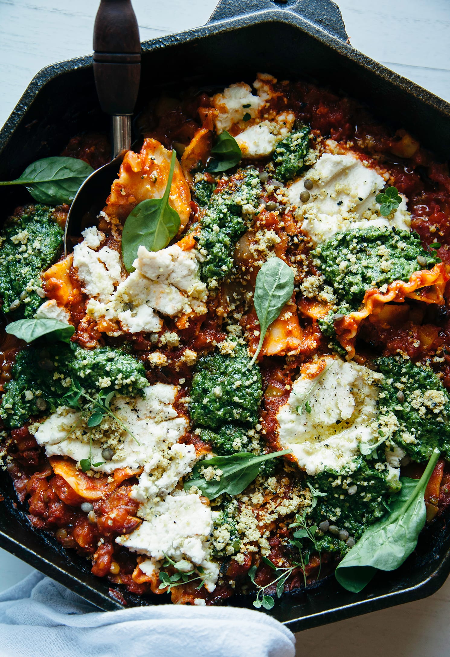 An overhead shot of a rustic, free form skillet lasagna topped with almond "ricotta" dollops, blobs of pesto, and spinach leaves. The dish is photographed in an octagonal cast iron pan.