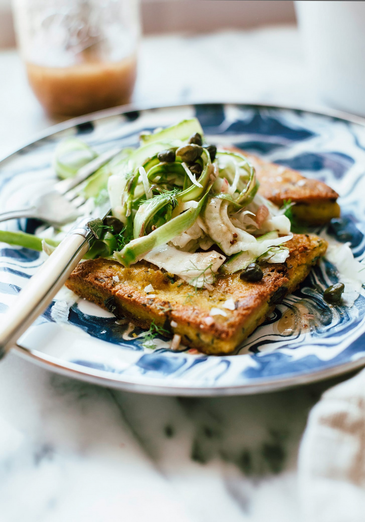 A 3/4 angle shot of two seared triangles of polenta on a marbled blue and white plate. The polenta is topped with a shaved vegetable salad.