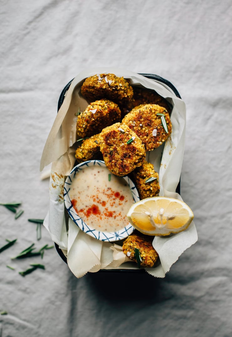 An overhead shot of golden grain and vegetable-based nuggets in a paper-lined loaf pan. A small bowl of creamy sauce and a lemon wedge is served on the side.