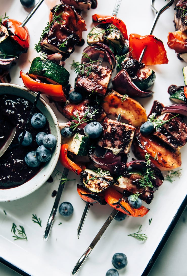 An overhead shot of grilled vegetable and tofu skewers in a white tray. The skewers are brushed with a blueberry bbq sauce.