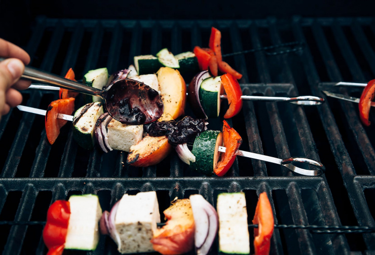 Image shows a hand basting grilled veg and tofu skewers with bbq sauce on the grill.