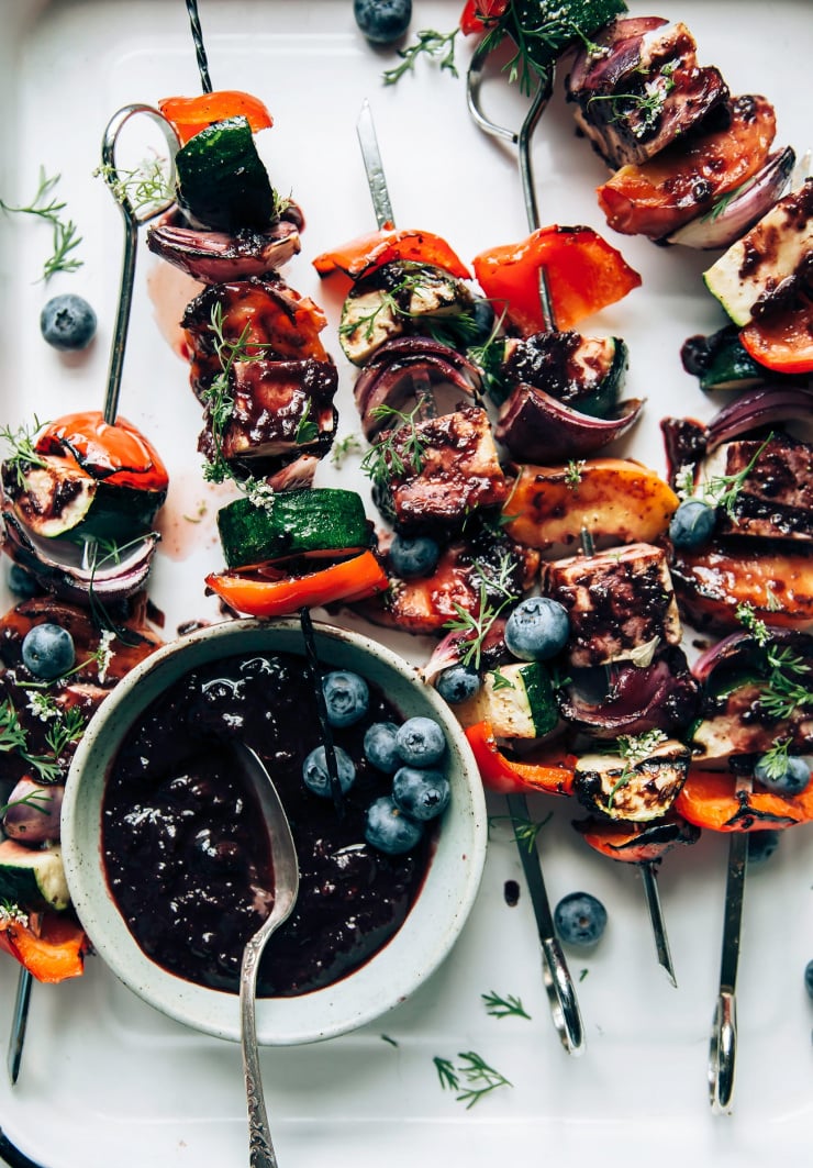 An overhead shot of grilled vegetable and tofu skewers in a white tray. The skewers are brushed with a blueberry bbq sauce.