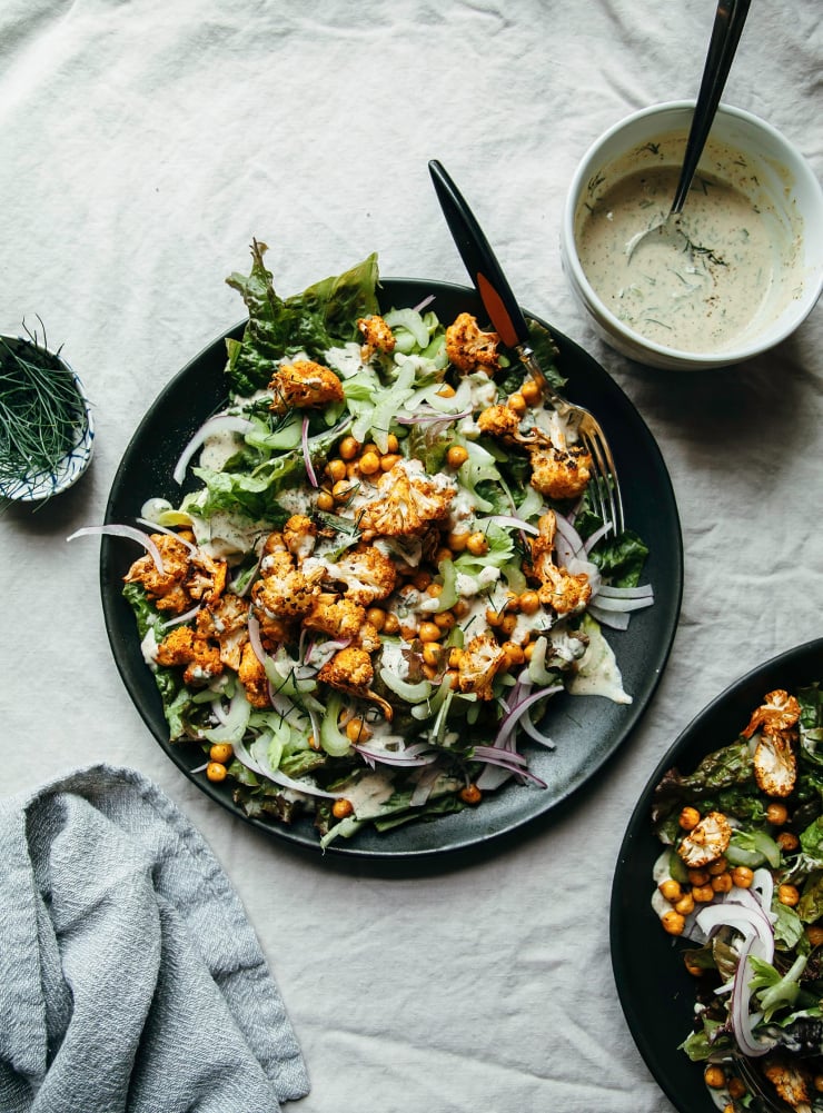 An overhead shot of a vegan Buffalo cauliflower salad on a matte black plate. A bowl of creamy dressing is nearby.