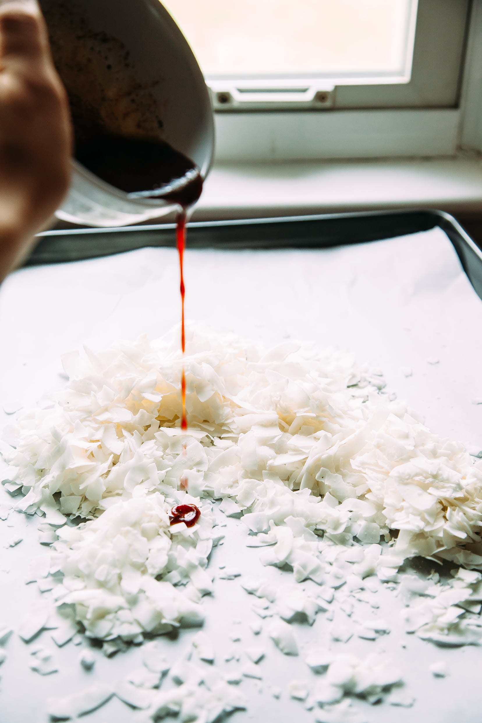 Image shows a hand pouring a deep red liquid over a sheet pan of coconut flakes.