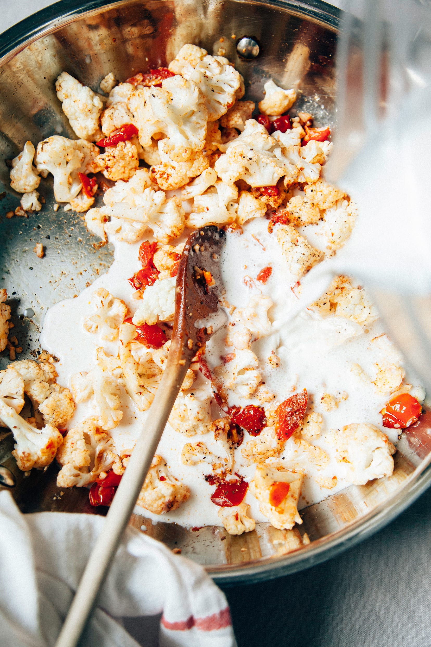 Image shows a creamy sauce being poured into a pan with tomatoes and cauliflower.