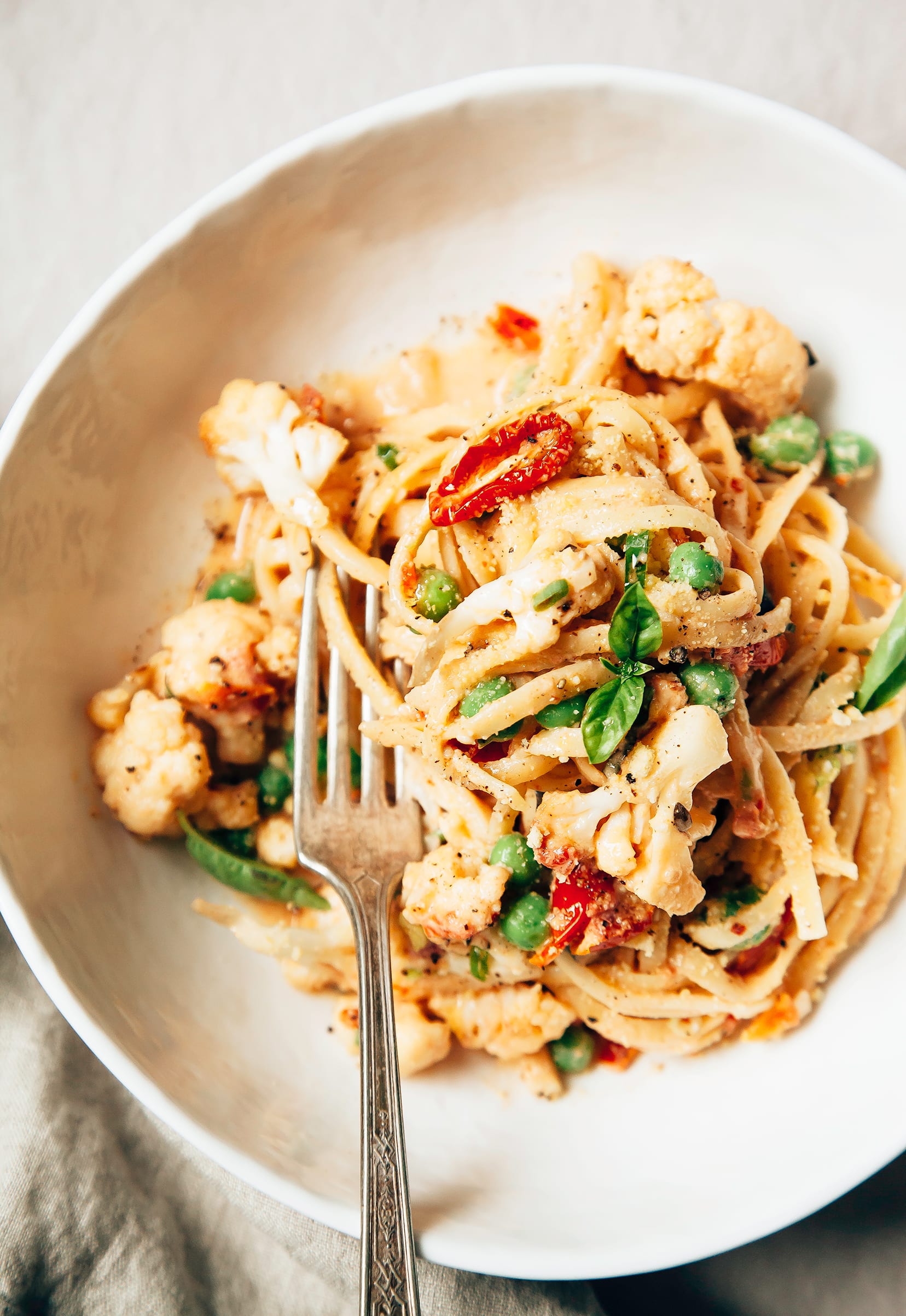 Image shows an individual serving of creamy cauliflower pasta with basil, peas, and tomatoes.