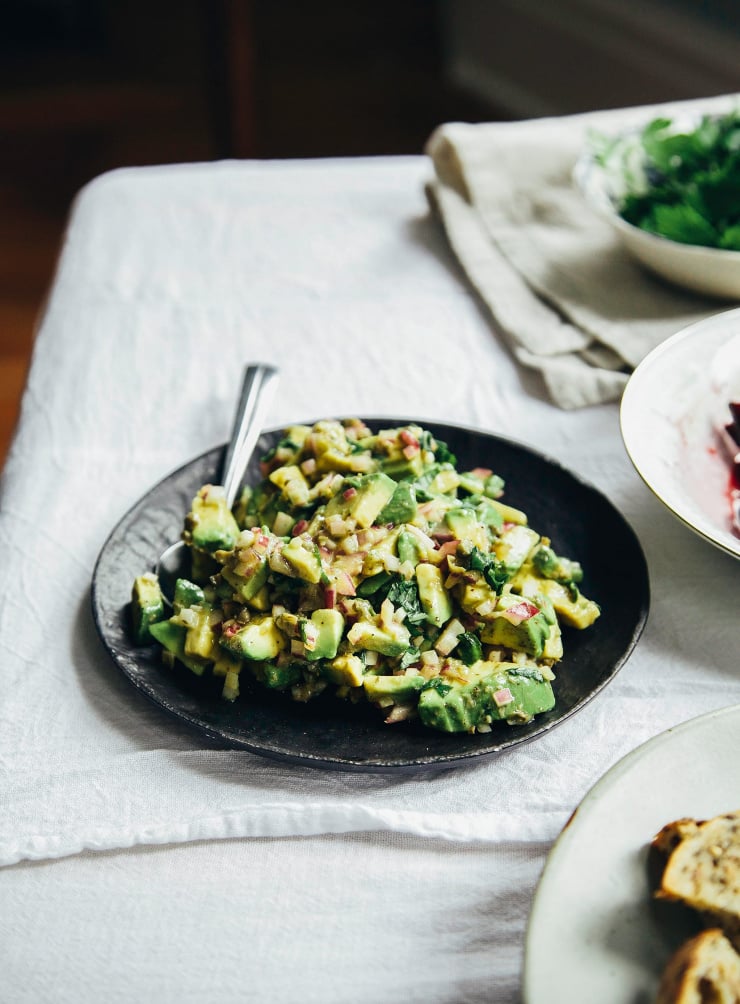 A 3/4 angle image of a plate with a diced avocado mixture on top. The plate is matte black and on top of a white linen tablecloth.