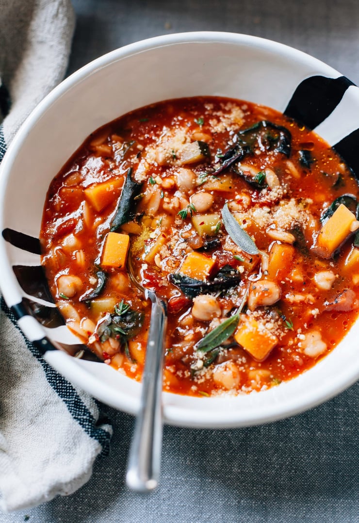 An up close, overhead shot of vegan butternut minestrone with sage, chickpeas, and chopped chard. The soup is deep red and chunky. It is plated in a wide white bowl with black streaks painted on to it. The minestrone is garnished with a vegan “Parmesan” style sprinkle.