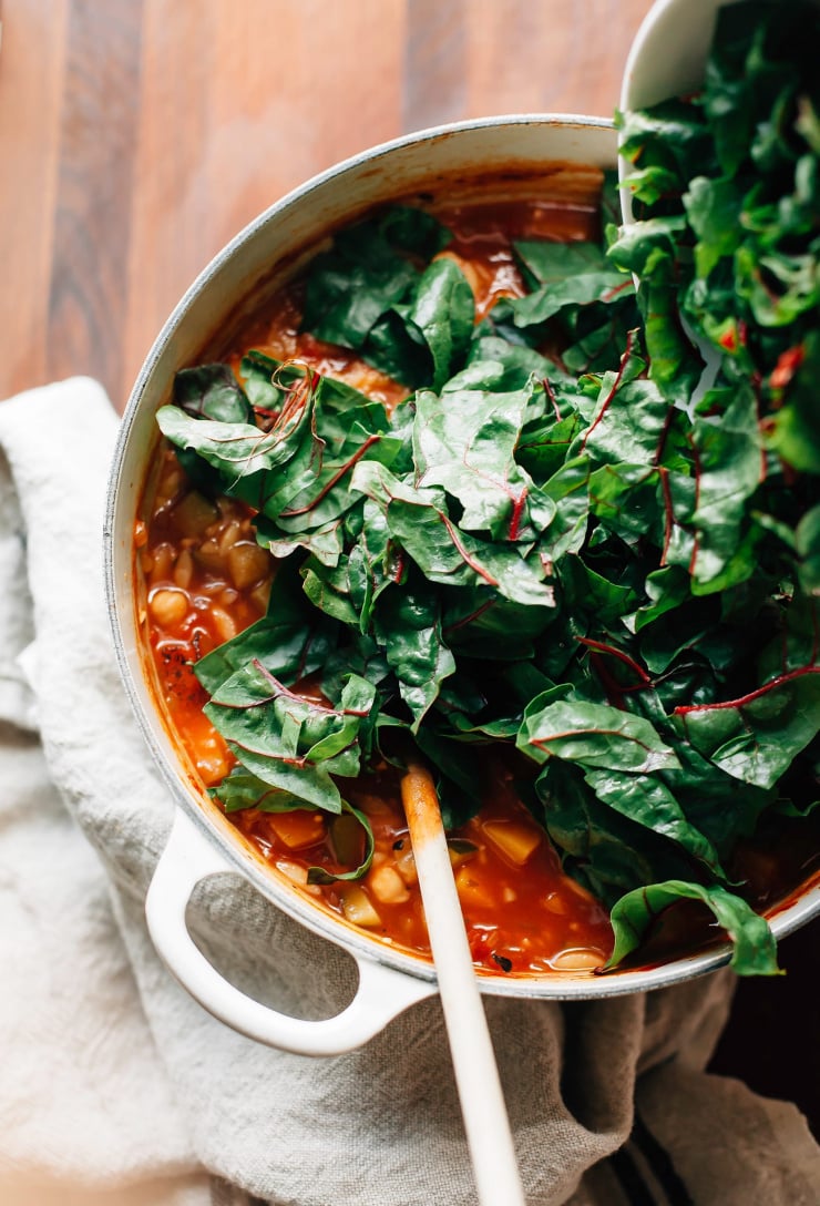 Chopped chard being added to a pot of red soup in an off-white Dutch oven.