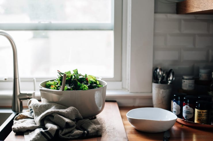 A head on shot of an off white Dutch oven with chard spilling out the top. The pot is set on a wooden cutting board in front of a kitchen window.