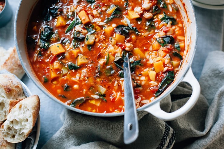 An overhead shot of butternut minestrone with chickpeas and chard in an off-white Dutch oven, set on top of a grey linen tablecloth. There is a bread basket nearby.
