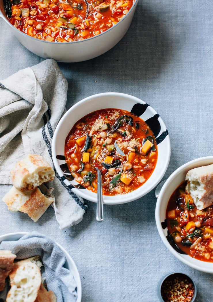 An overhead shot of butternut minestrone with chickpeas and chard in white bowls, set on top of a grey linen tablecloth. There is a bread basket nearby. The bowls are garnished with a sprinkle of nut-based “Parmesan”