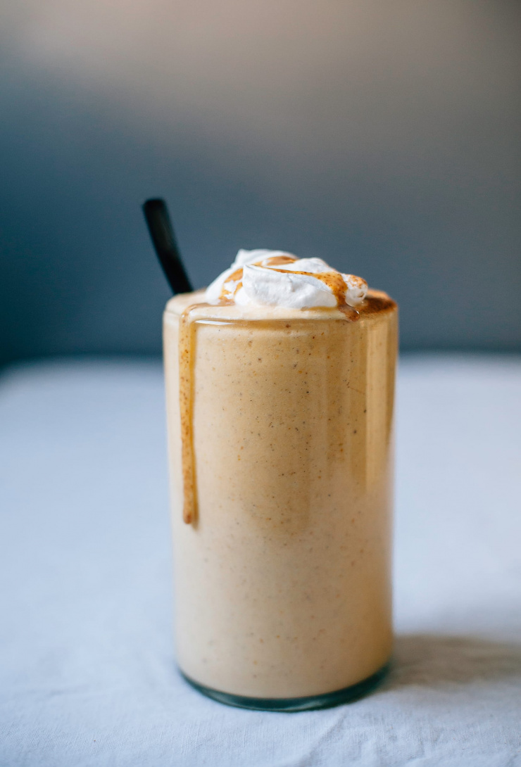 An up close, head-on shot of a light orange, very creamy smoothie in a clear glass that is garnished with bright white coconut cream and a drizzle of almond butter. A spoon is sticking out of the glass.