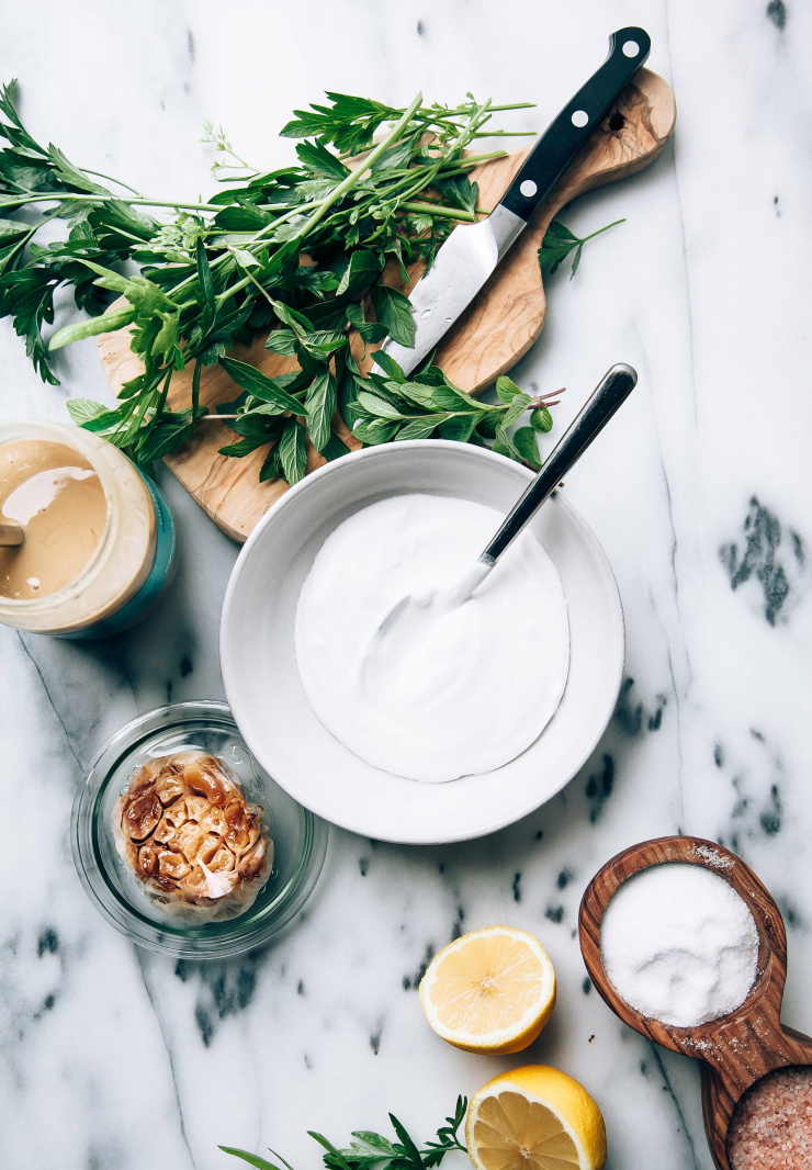An overhead image shows ingredients for a roasted garlic tahini yogurt dip on a marble background.
