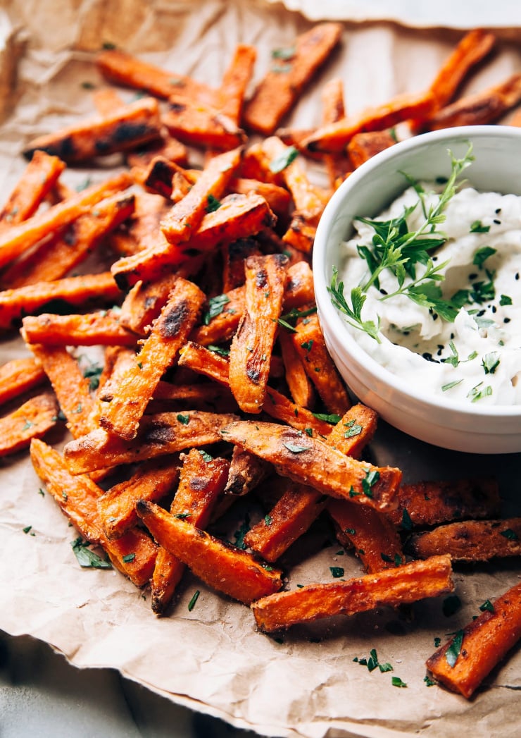 A close up shot of crispy, slightly browned sweet potato fries on crumpled up brown paper. There is a creamy white dip in a bowl to the side.