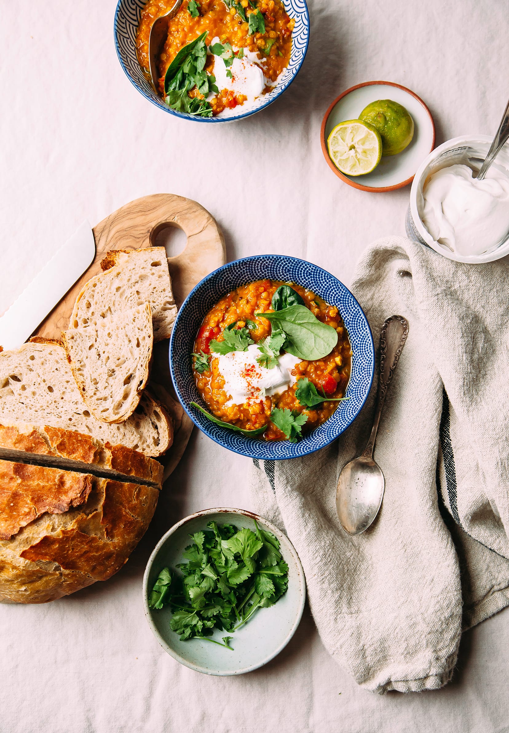 An overhead shot of bowls of a deep orange lentil soup with spinach, along with bread served on the side.