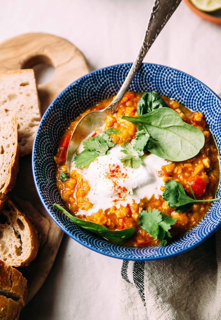 Golden lentil soup with coconut spinach and lime, photographed from overhead in a deep blue bowl, garnished with a dollop of coconut yogurt. There is a wood board with sliced bread nearby. All is photographed on a beige linen tablecloth.