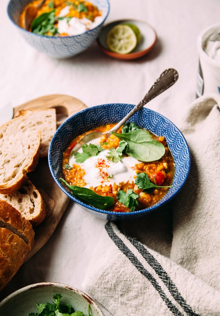 Golden lentil soup with coconut spinach and lime, photographed from a 3/4 angle in a deep blue bowl, garnished with a dollop of coconut yogurt. There is a wood board with sliced bread nearby. All is photographed on a beige linen tablecloth.